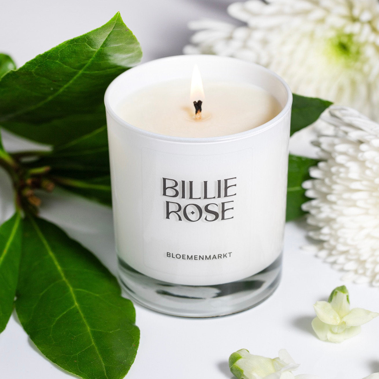 White candle in a glass holder with 'Billie Rose' branding, surrounded by green leaves and white flowers.