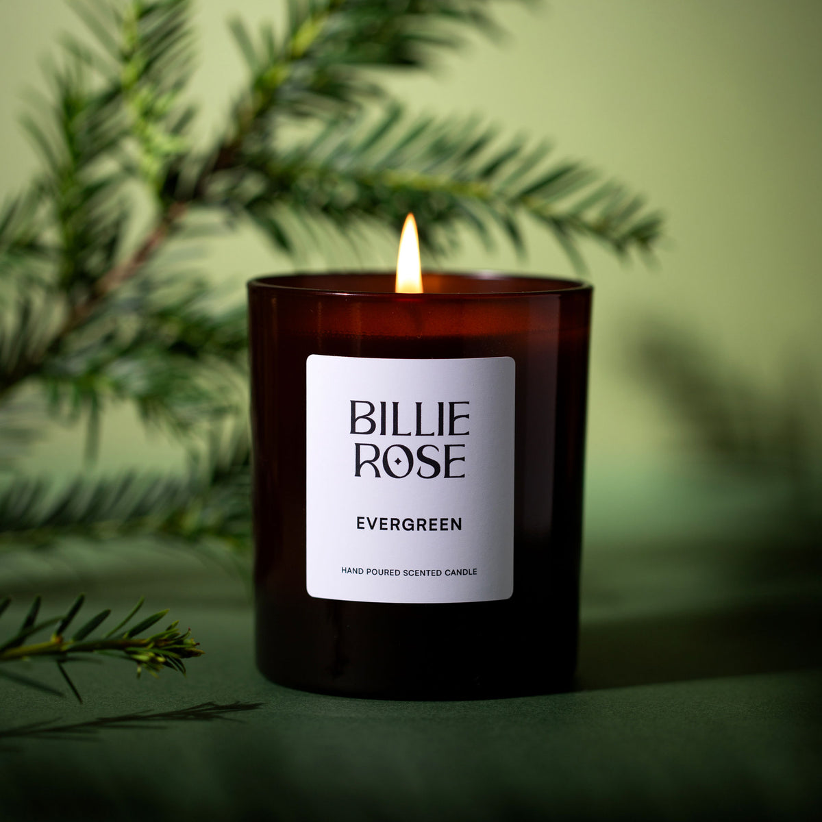 A lit Billie Rose Evergreen hand poured scented candle in a dark amber glass jar, styled with pine branches against a soft olive green background.