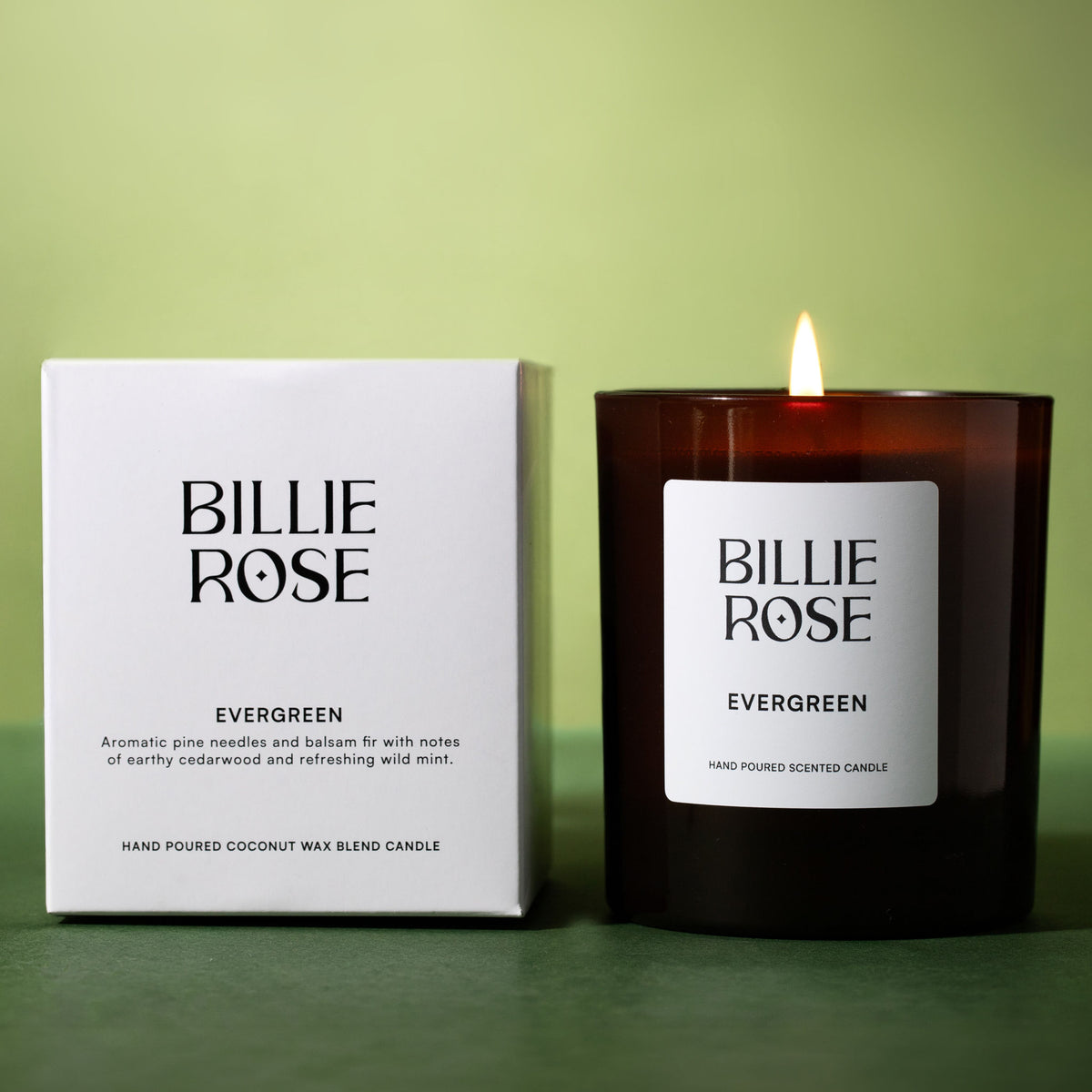 A Billie Rose Evergreen scented candle in an amber glass jar next to its white packaging box, set against a soft green background with the candle lit.