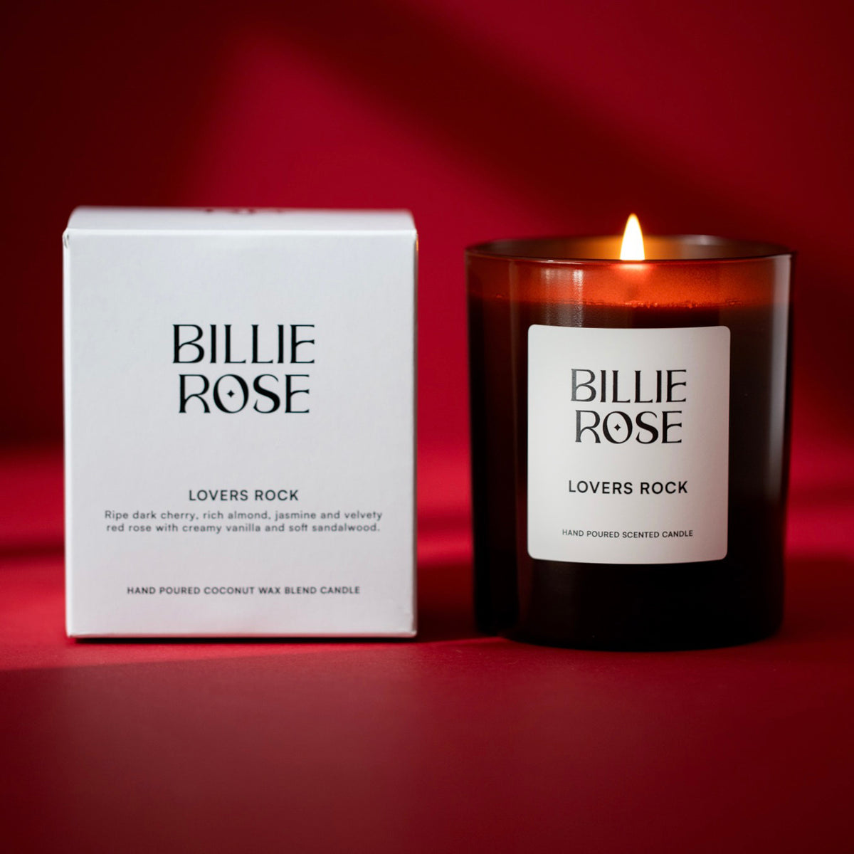 A lit Billie Rose 'Lovers Rock' scented candle in a dark amber glass jar sits next to its white packaging box on a deep red background.