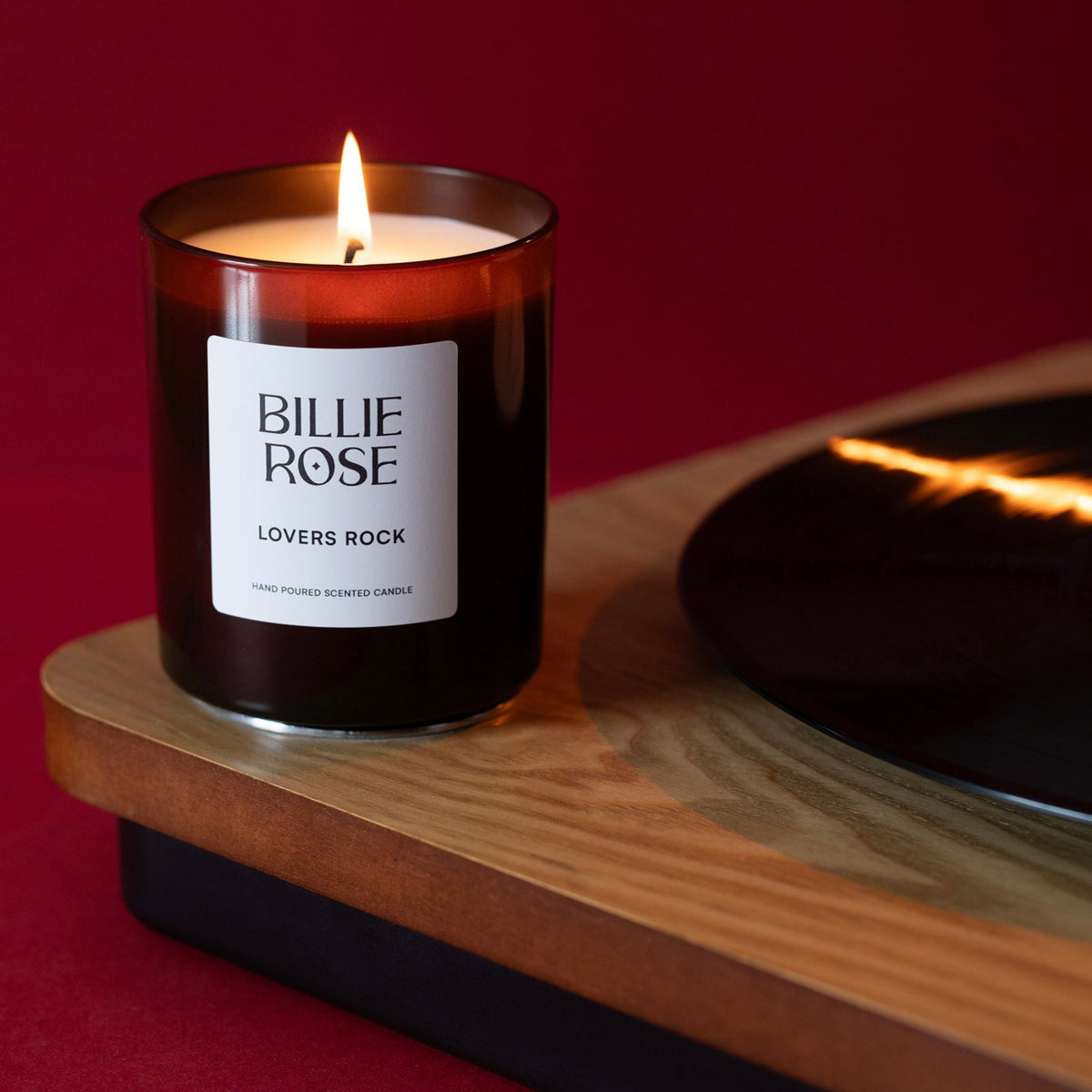 A Billie Rose 'Lovers Rock' scented candle in an amber glass jar, lit and sitting on a record player against a deep red background.