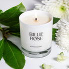 White candle in a glass holder with 'Billie Rose' branding, surrounded by green leaves and white flowers.