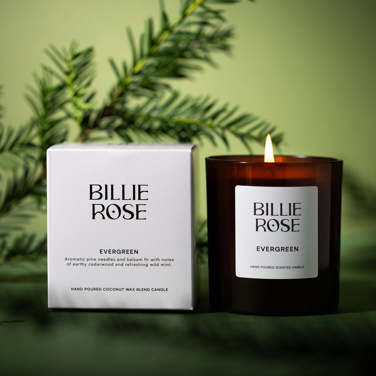 A Billie Rose Evergreen scented candle in a dark amber glass jar next to its white packaging box, set against a soft olive green background with blurred pine branches.