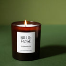 A lit Billie Rose Evergreen scented candle in an amber glass jar, set against a solid dark green background.