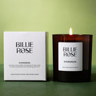 A Billie Rose Evergreen scented candle in an amber glass jar next to its white packaging box, set against a soft green background with the candle lit.