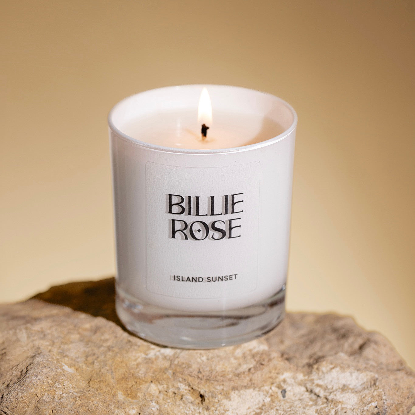 Candle in a glass container labelled 'Billie Rose' on a stone surface with a warm background