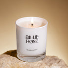 Candle in a glass container labelled 'Billie Rose' on a stone surface with a warm background