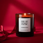 A lit Billie Rose 'Lovers Rock' scented candle in a dark amber glass jar on a deep red background with piece of red ribbon to the left.