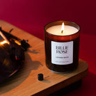 A lit Billie Rose 'Lovers Rock' scented candle in a dark amber glass jar, styled on a wooden turntable next to a vinyl record against a deep red background.