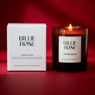 A lit Billie Rose 'Lovers Rock' scented candle in a dark amber glass jar sits next to its white packaging box on a deep red background.
