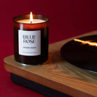 A Billie Rose 'Lovers Rock' scented candle in an amber glass jar, lit and sitting on a record player against a deep red background.