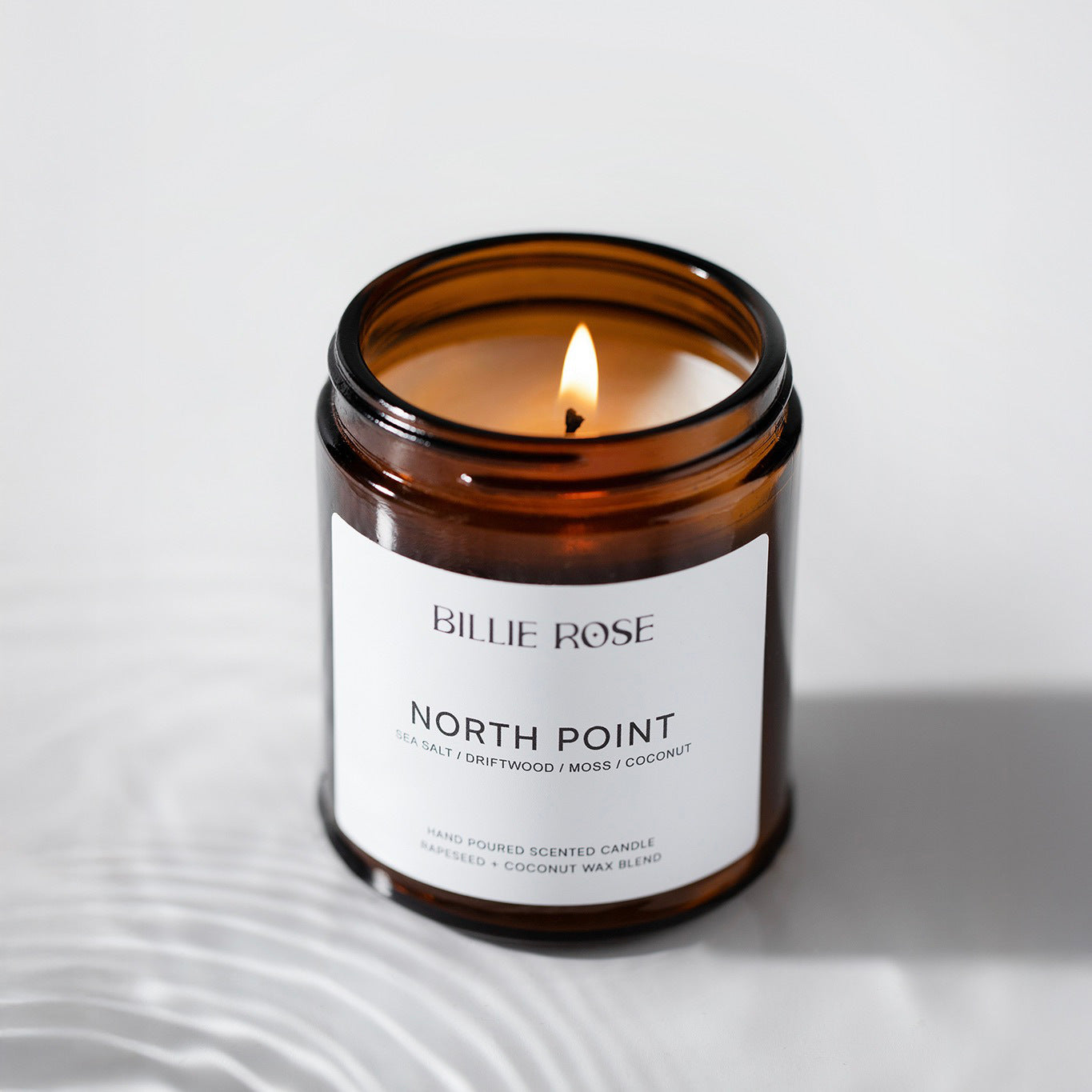 Lit candle in a brown glass jar with 'Billie Rose' label on a light gray background