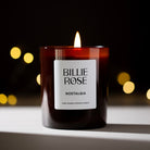 A lit Billie Rose Nostalgia scented candle in a dark amber glass jar, set against a dark background with warm, out-of-focus bokeh lights.