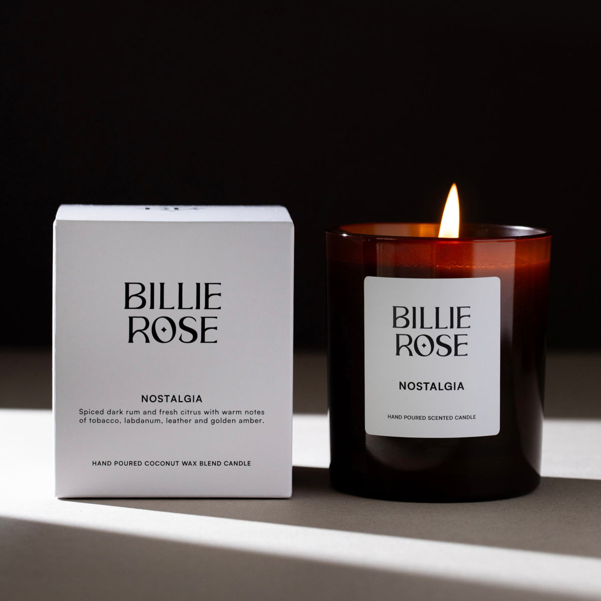 Billie Rose Nostalgia hand-poured rapeseed and coconut wax candle shown with its white product box and a lit amber glass jar candle on a dark surface against a black background.