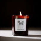 A lit Billie Rose Nostalgia scented candle in an amber glass jar, set against a dark background with a warm flame burning at the top.