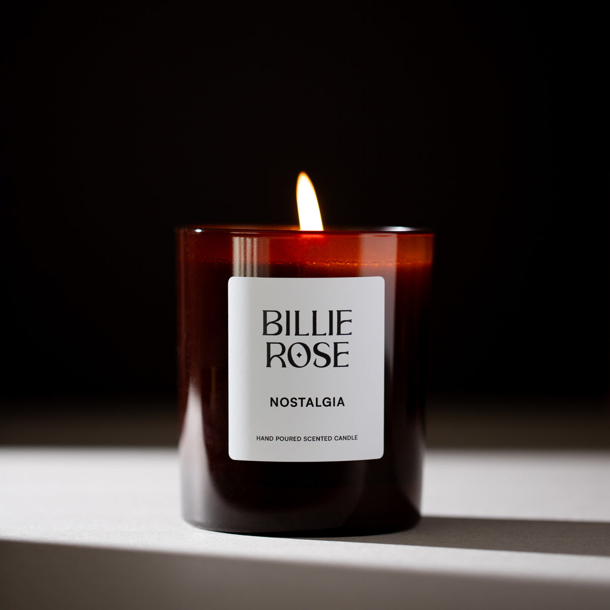 A lit Billie Rose Nostalgia scented candle in an amber glass jar, set against a dark background with a warm flame burning at the top.