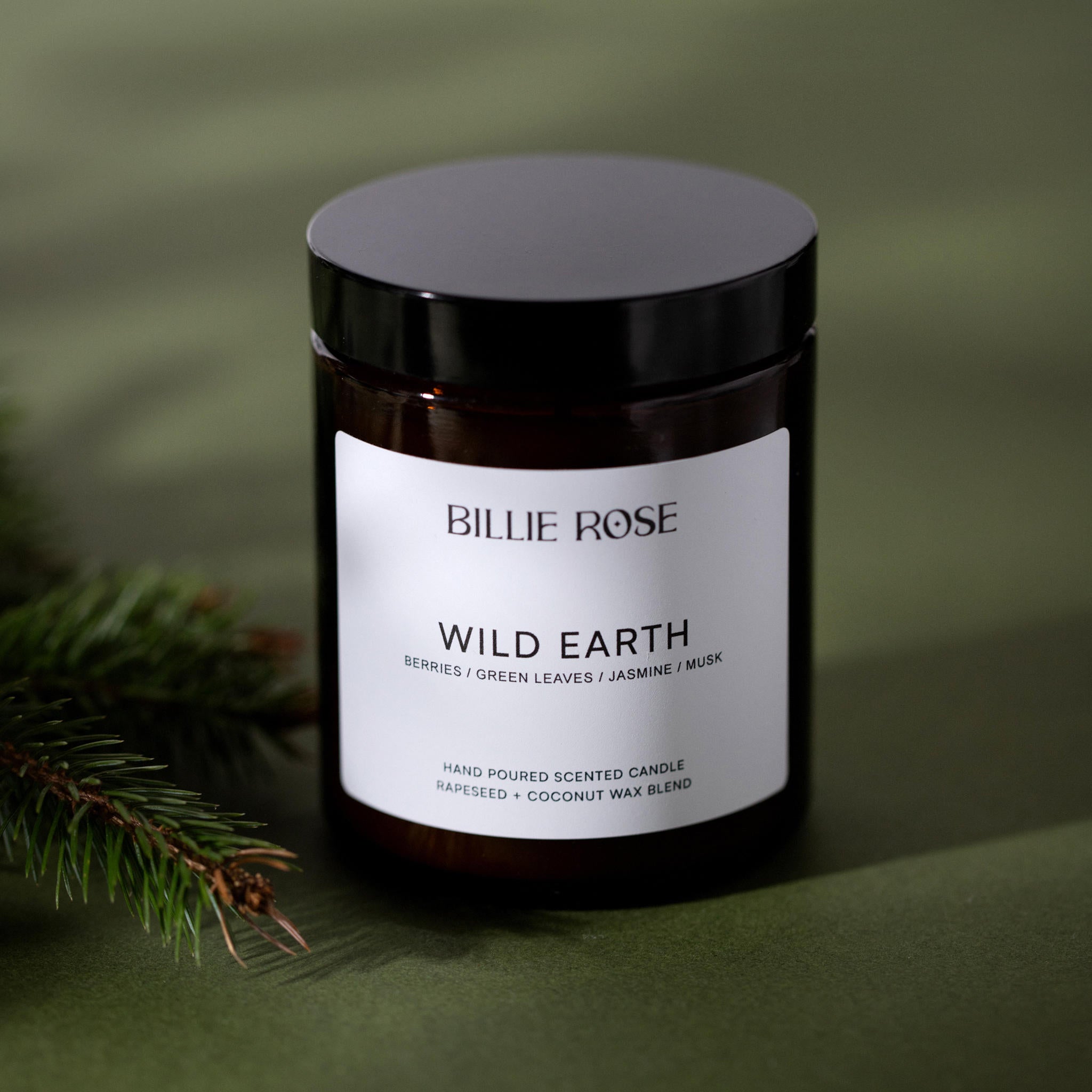 Candle jar labeled 'Billie Rose Wild Earth' on a green background with a pine branch.