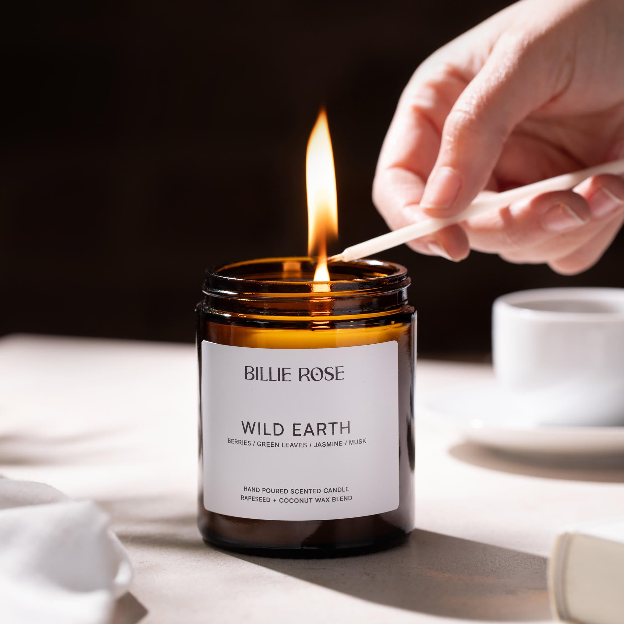 Candle labelled 'Billie Rose Wild Earth' being lit with a match on a neutral background.