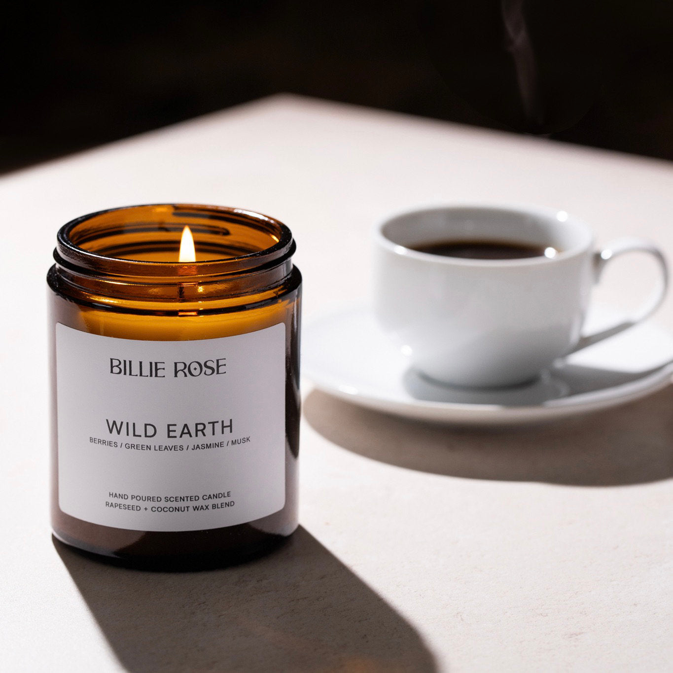 Billie Rose Wild Earth candle with a lit flame next to a cup of coffee on a white surface.