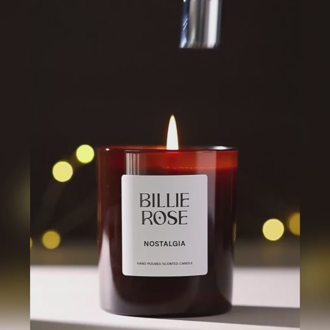 video of Billie Rose Nostalgia candle being put out with a candle snuffer and wisps of smoke rising against a dark brown background with blurred bokeh lights